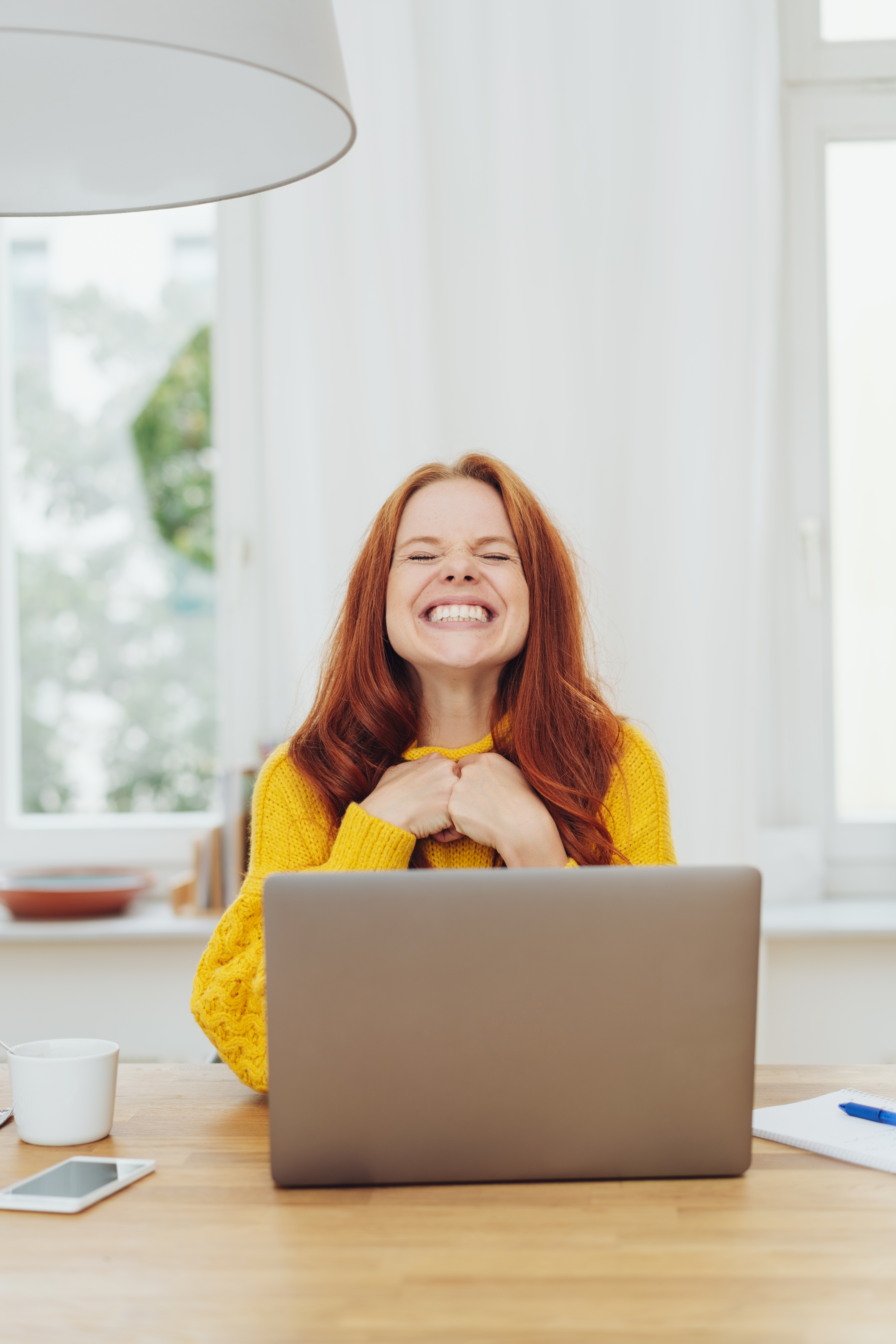 Excited young businesswoman screwing up her eyes and grinning with a beaming toothy smile as she works at a laptop